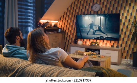 Happy couple sitting on sofa, watching action movie on TV or criminal blockbuster on streaming service, talking and discussing acting, eating snacks, pizza, chips. Spouses resting at home in evening. - Shutterstock ID 2213917389