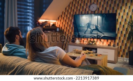 Happy couple sitting on sofa, watching action movie on TV or criminal blockbuster on streaming service, talking and discussing acting, eating snacks, pizza, chips. Spouses resting at home in evening.