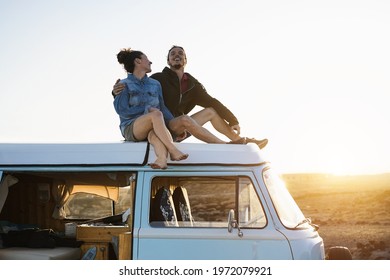 Happy couple sitting on top of minivan roof at sunset - Young people having fun on summer vacation traveling around the world - Love and holiday concept - Focus on faces