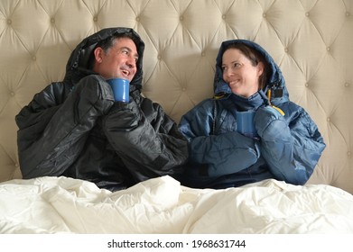 Happy Couple Sitting Side By Side Wearing A Very Warm Clothing And Covered With A Blanket, Sitting On A  Double Bed Looking At Each Other, Having A Hot Drink In Bedroom  On A Cold Winter Day.