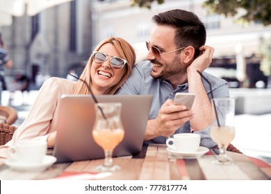 Happy couple sitting in street cafe together and using laptop and phone.