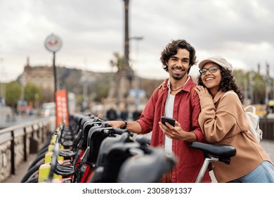 A happy couple is smiling at the camera while standing next to rental bikes on the street with a phone.