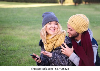 happy couple smiling together in the park. pretty girl and boy in autumn winter clothes out on a green park girl with cellphone.