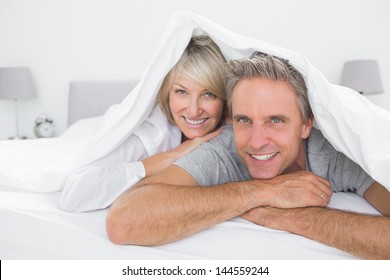 Happy Couple Smiling Under The Covers At The Camera At Home In Bed