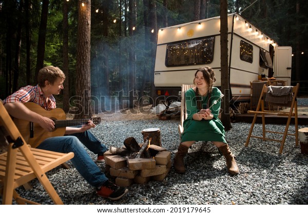 happy couple (smiling woman and man playing\
guitar) sits close to campfire and trailer (house on wheel) in\
forest summer camping