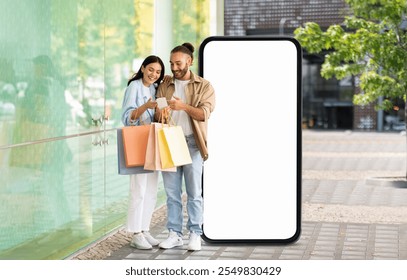 A happy couple stands outside a store next to huge phone with blank screen, looking at their phone and smiling. They hold colorful shopping bags, enjoying their day together under clear skies.