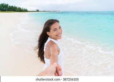 Happy couple summer vacation travel - Asian woman on romantic honeymoon beach holiday holding hand of boyfriend looking at camera. Man following girlfriend walking. POV.