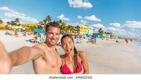 Happy Couple Taking Fun Selfie On Florida Beach On Travel Summer Vacation. Suntan Man And Asian Bikini Woman In Bikini Using Phone Laughing POV. Interracial Group Portrait On Fort Myers Beach Holiday.