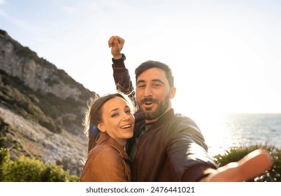 Happy couple taking a selfie on a sunny day by the sea. They are smiling and embracing with a scenic cliff and the ocean in the background, capturing a moment of joy and adventure. - Powered by Shutterstock