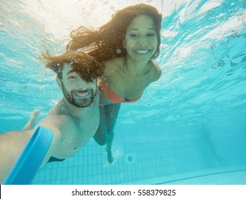 Happy couple taking selfie under the water in swimming pool resort with special photo video camera - Young people having fun in summer vacation - Holidays concept - Focus on man face - Warm filter - Powered by Shutterstock