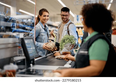 Happy couple talking to cahier while putting groceries on checkout counter in the supermarket.