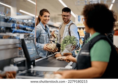 Happy couple talking to cahier while putting groceries on checkout counter in the supermarket.