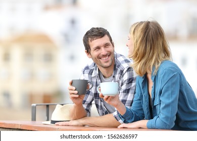 Happy couple talking holding coffee mugs standing in a balcony outdoors in a rural town