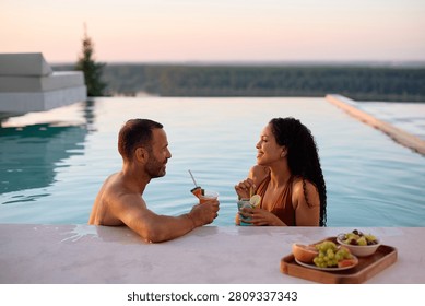 Happy couple talking while drinking cocktails in infinity pool in summer. Copy space. 