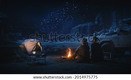 Happy Couple Tent Camping in the Canyon, Sitting by Campfire Watching Night Sky with Milky Way Full of Bright Stars. Two Travelers In Love On a Romantic Vacation Trip. Back View Shot