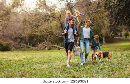 Happy Couple With Their Two Children And Dog Walking In Park. Man Carrying Little Girl With Kite On Shoulders And Woman Carrying A Picnic Basket With Son And Pet Dog.