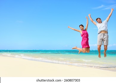 Happy Couple Tourists Jumping On Beach Vacations. Travel Concept Of Young Couple Cheering For Summer Holidays Showing Success, Happiness, And Joy On Perfect White Sand Tropical Beach Under The Sun.