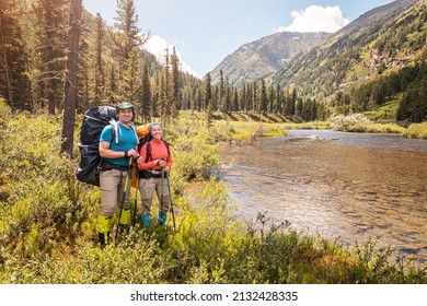 Happy couple travelers with trekking poles and a large backpack walks along a trail in a national park with a mountain river in the background. - Shutterstock ID 2132428335