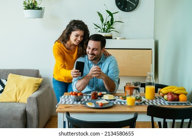 Happy Couple Using Mobile Phone Together While Eating Breakfast at Home. 
Smiling girlfriend showing phone to her boyfriend while having healthy breakfast in the dining room. - Powered by Shutterstock