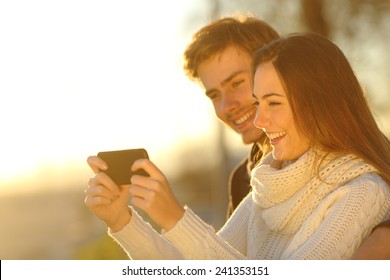 Happy Couple Watching Media Videos In A Smart Phone At Sunset On The Beach