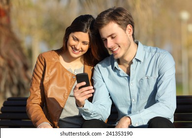 Happy Couple Watching A Smart Phone Sitting On A Bench In A Park