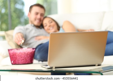 Happy couple watching tv on line in a laptop and eating popcorn lying on a comfortable sofa at home