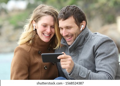 Happy Couple Watching Videos On Smart Phone Sharing Earbuds On The Beach In Winter