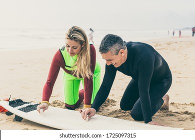 Happy couple waxing surfboard on beach. Cheerful man and woman in wetsuits sitting on sand and waxing surfboard at ocean coast. Surfing concept - Powered by Shutterstock