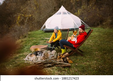 Happy Couple Of Weekend Tourists Sit On Sun Chairs In Front Of Campfire At Glamorous Camping Site Near Glamping Tent. Weekend Getaway Destination. Staycation Adventure