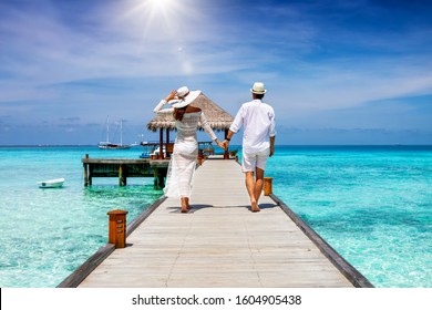 A happy couple in white summer clothing on vacation walks along a wooden pier over tropical, turquoise ocean in the Maldives, Indian Ocean