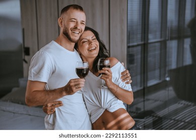 Happy couple in white t-shirts holding glass of wine smiling at home. Bearded caucasian man embracing wife laughing celebrates anniversary at hotel. Honeymoon. Excited newlyweds on vacations. - Powered by Shutterstock