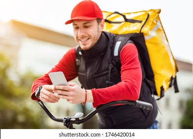 Happy Courier Using Phone Delivering Food From Restaurants On Bike Waiting For Meal Order Outdoors. Delivery Service. Selective Focus
