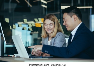 Happy Coworkers Discuss Project Strategy By Looking And Pointing At Laptop Pc Computer Monitor Screen. Multiethnic Business Team In The Office. Confident Mature Asian Man Explaining Young Female