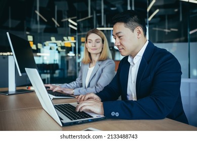 Happy Coworkers Discuss Project Strategy By Looking And Pointing At Laptop Pc Computer Monitor Screen. Multiethnic Business Team In The Office. Confident Mature Asian Man Explaining Young Female