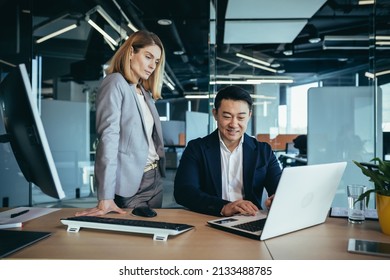 Happy Coworkers Discuss Project Strategy By Looking And Pointing At Laptop Pc Computer Monitor Screen. Multiethnic Business Team In The Office. Confident Mature Asian Man Explaining Young Female