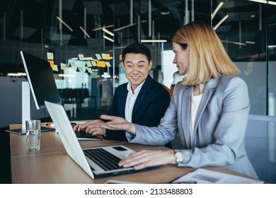 Happy Coworkers Discuss Project Strategy By Looking And Pointing At Laptop Pc Computer Monitor Screen. Multiethnic Business Team In The Office. Confident Mature Asian Man Explaining Young Female