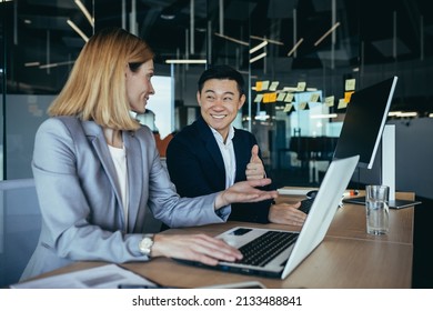 Happy Coworkers Discuss Project Strategy By Looking And Pointing At Laptop Pc Computer Monitor Screen. Multiethnic Business Team In The Office. Confident Mature Asian Man Explaining Young Female