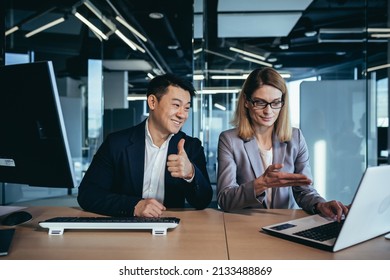 Happy Coworkers Discuss Project Strategy By Looking And Pointing At Laptop Pc Computer Monitor Screen. Multiethnic Business Team In The Office. Confident Mature Asian Man Explaining Young Female