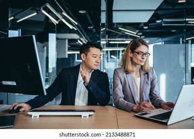 Happy Coworkers Discuss Project Strategy By Looking And Pointing At Laptop Pc Computer Monitor Screen. Multiethnic Business Team In The Office. Confident Mature Asian Man Explaining Young Female