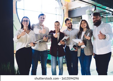 Happy coworkers in formal and casual wear holding files and showing thumbs up while looking at camera in office hall - Shutterstock ID 1613594296