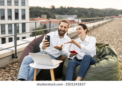 Happy coworkers sitting on rooftop enjoying break using smartphone and laptop. Smiling man and woman in casual attire working outdoors feeling relaxed and joyful. Fun and creative working environment. - Powered by Shutterstock