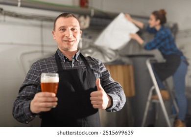 Happy craft brewery owner inviting to tasting fresh amber beer of own production, showing thumbs up while holding out glass of drink - Powered by Shutterstock