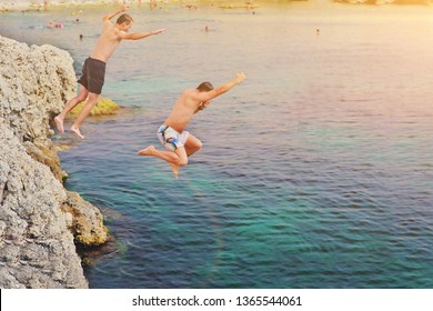 Happy Crazy Friends Jumping Inside Natural Sea Pool From Rock At Sunset. Young Fearless Two Guys People Having Fun Diving Inside Ocean Water From Cliff. Summer Lifestyle And Adventure Vacation Concept