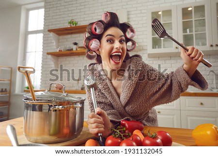 Happy crazy goofy young woman having fun while making healthy vegetable soup at home. Funny positive excited housewife in hair rollers and bathrobe standing at kitchen table, cooking meal and laughing