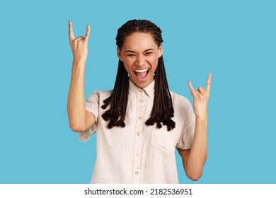 Happy Crazy Woman With Dreadlocks Showing Rock And Roll Gesture, Heavy Metal Sign, Enjoys Favorite Music On Party, Has Squints Face, Exclaims From Joy. Indoor Studio Shot Isolated On Blue Background.