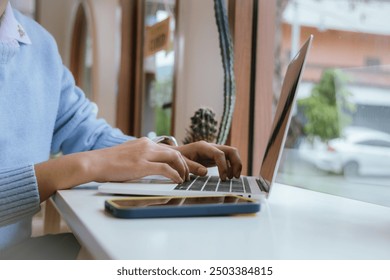 A happy, creative businesswoman sits at a wooden desk in a cozy cafe, using a laptop and smartphone for online communication. She types on the keyboard, reflecting a modern, connected lifestyle. - Powered by Shutterstock