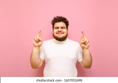 Happy curly fat man stands on a pink background and makes a wish with crossed fingers on a pink background. Cheerful overweight boy crossed fingers with closed eyes, wearing white t-shirt, isolated.