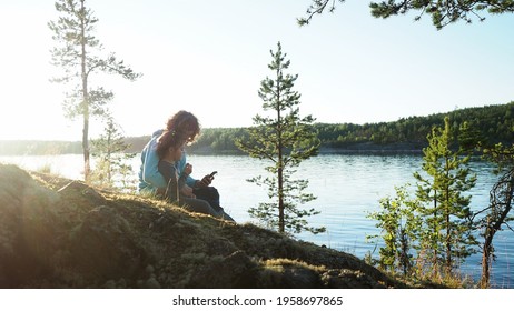 Happy Curly Hair Mother With Little Daughter Sitting By Lake During Sunset On Spring Camping Vacation And Browsing Internet On Mobile Phone Device Watching Video. Concept Of Nature Quality Family Time