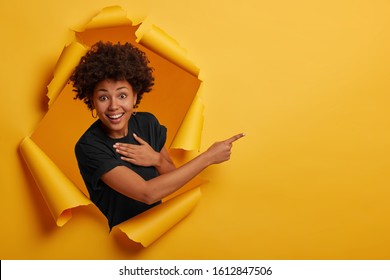 Happy curly haired African American woman laughs positively, points aside on copy space, wears black t shirt, shows cool advertisement or promotion, isolated over yellow background. Torn paper