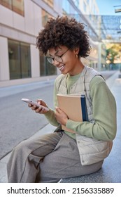 Happy curly haired woman uses mobile application for browsing media content reads receieved message holds textbooks wears jumper vest and trousers sits outdoors during sunny day. Technology concept